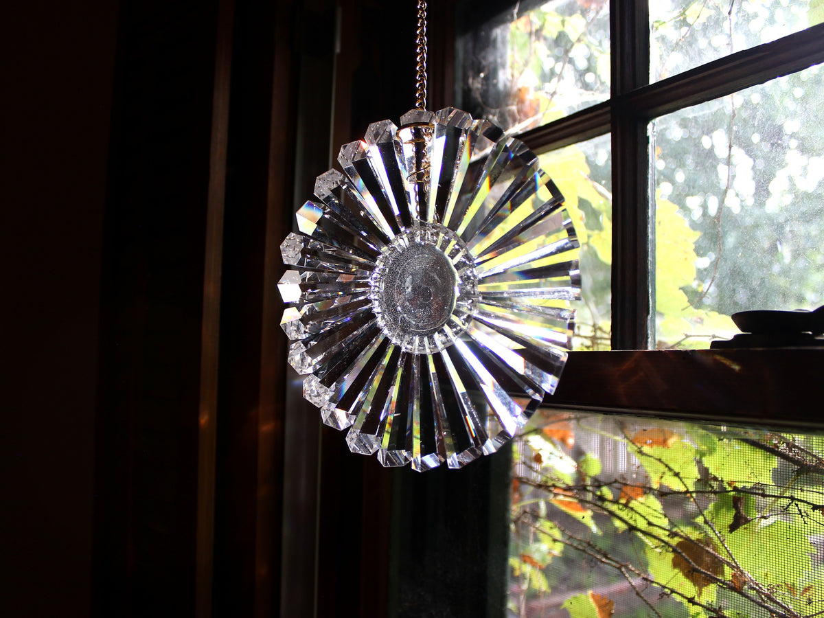 A clear faceted lead crystal sun catcher or suncatcher hanging from a chain in front of a window, with light rays passing through the circular, sunburst-shaped design.