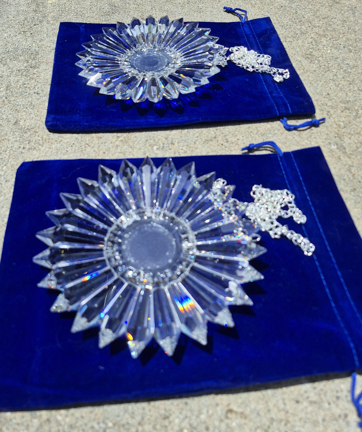 Two clear faceted crystal prism ornaments with silver-toned chains, displayed on dark blue velvet drawstring pouches on a sandy surface.