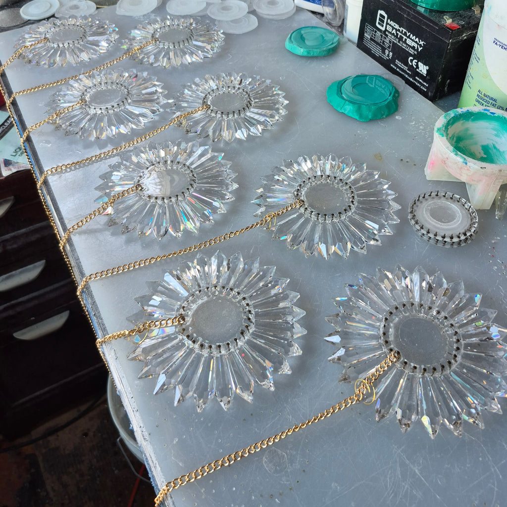 Eight clear faceted prism suncatcher rainbow makers with gold chains are laid out on a gray workbench, surrounded by craft supplies including green paint lids, a battery, and a paint cup.