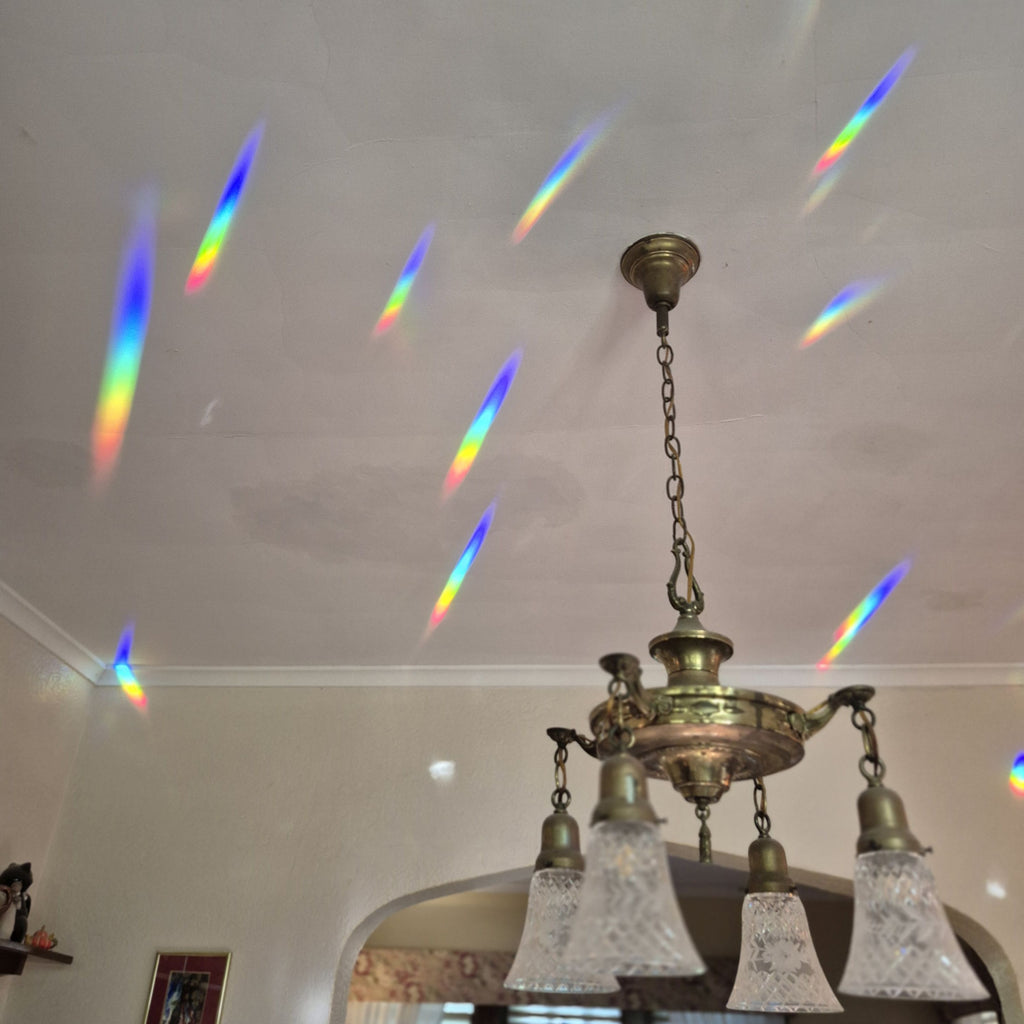 A brass multi-light chandelier hanging from a white ceiling, with colorful rainbow light beams projected across the ceiling from the prism suncatcher in the adjacent window.