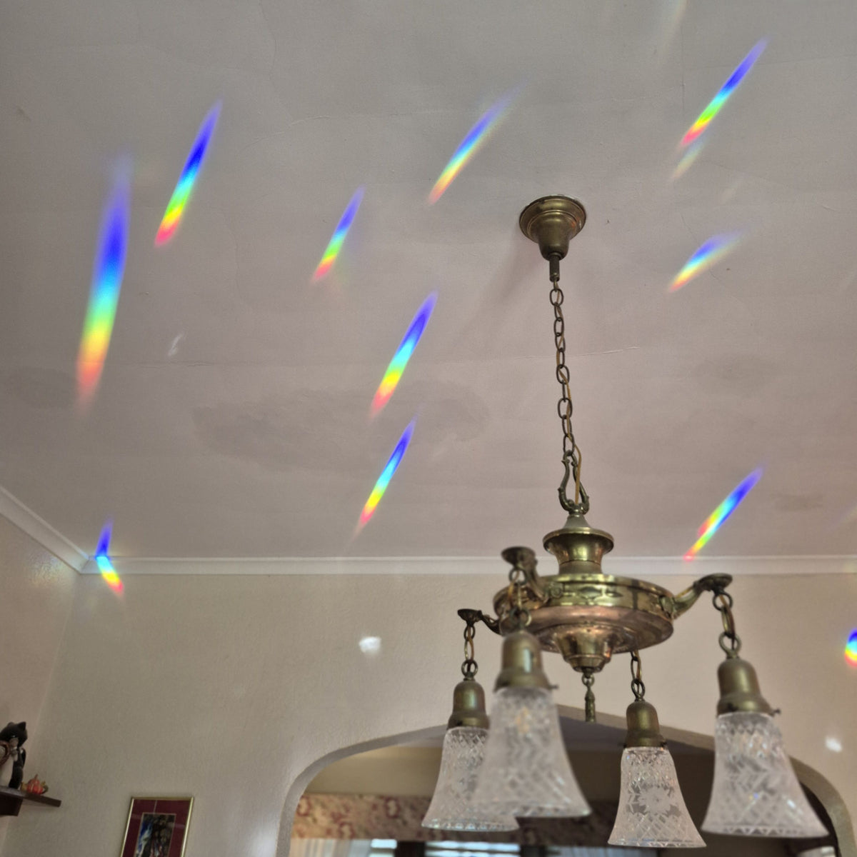 A brass multi-light chandelier hanging from a white ceiling, with colorful rainbow light beams projected across the ceiling from the prism suncatcher in the adjacent window.