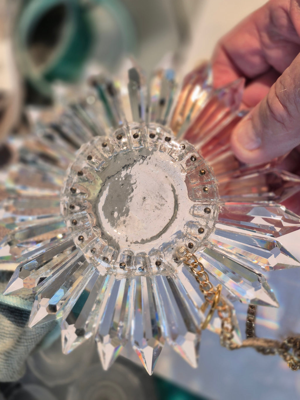 A hand holding a faceted clear crystal sunburst ornament with a gold-tone chain, shown close-up against a softly blurred background.