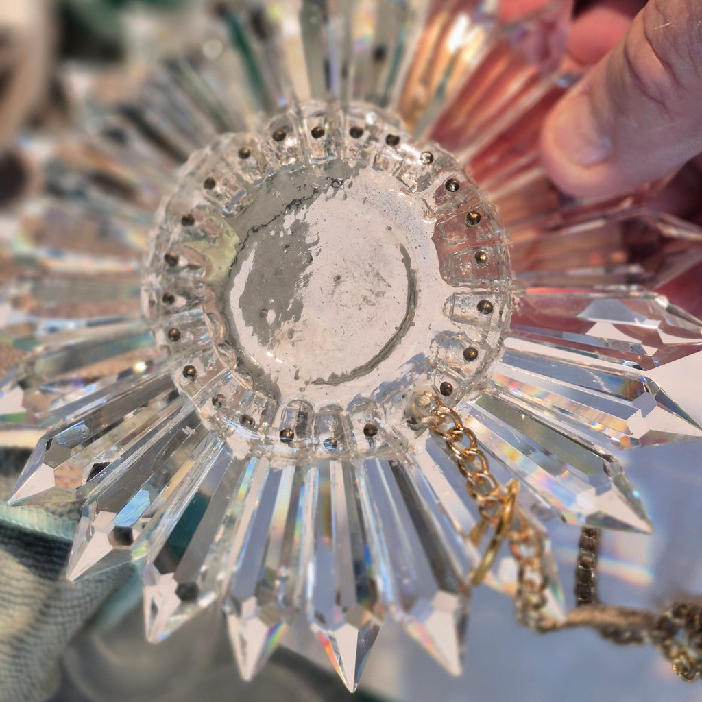 A hand holding a faceted clear crystal sunburst ornament with a gold-tone chain, shown close-up against a softly blurred background.