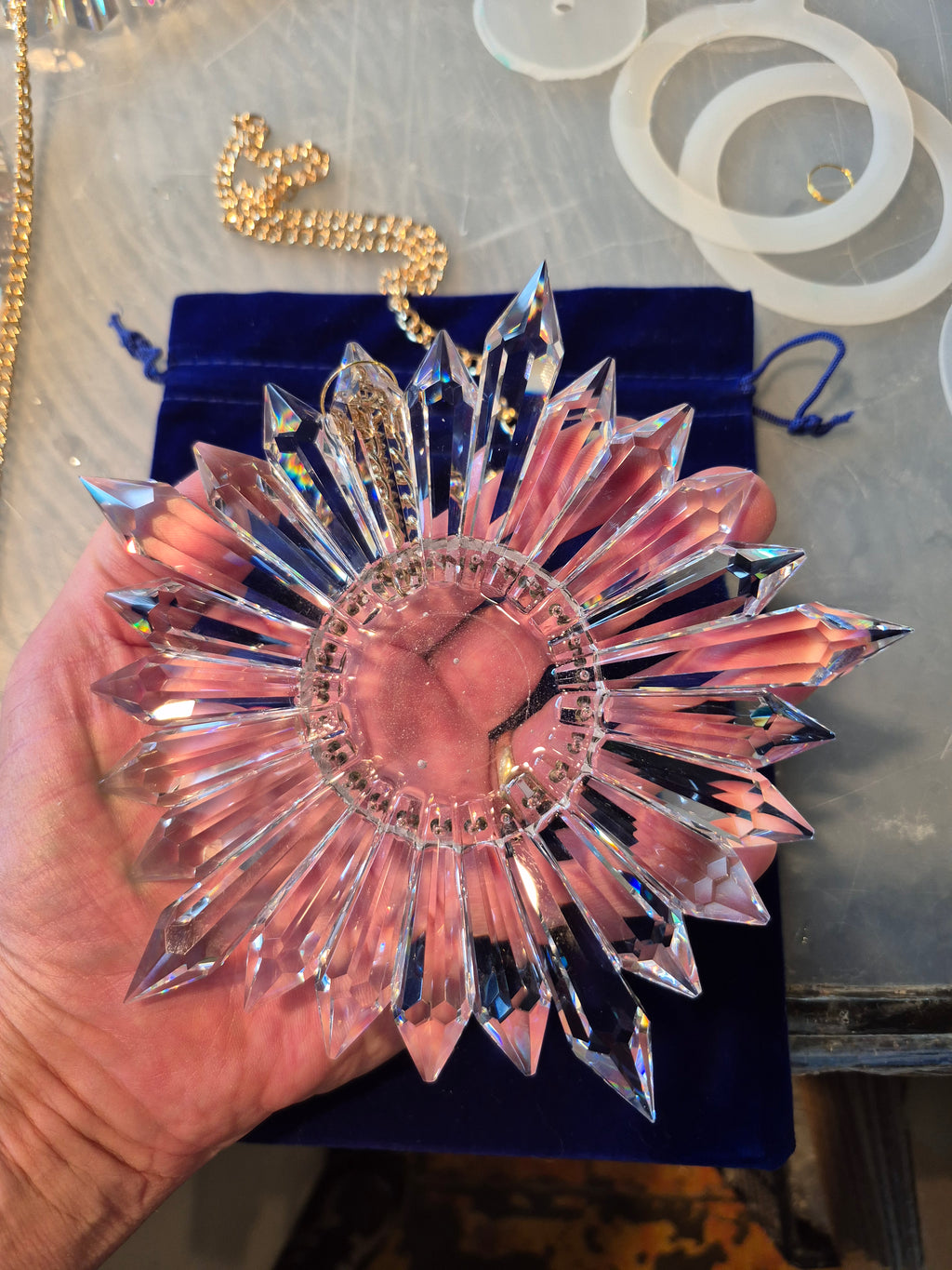 A hand holding a faceted clear crystal sunburst ornament, with a dark blue velvet pouch underneath on a tabletop. The crystal ornament has multiple pointed rays radiating from a circular center.