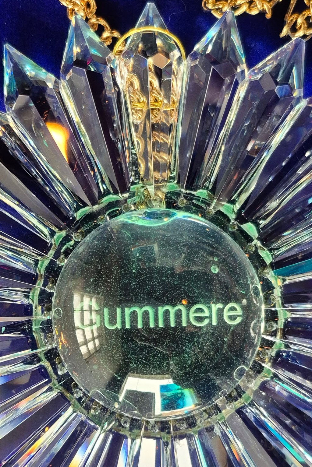 A close-up of a faceted clear crystal sunburst ornament or pendant with a central circular section printed with the word 'Gummere' in green, set against a dark blue background.