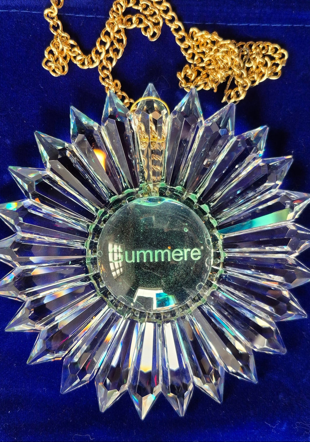 A faceted clear lead crystal prism suncatcher with a gold chain, displayed on a blue velvet pouch. The pendant features a central glass dome engraved.
