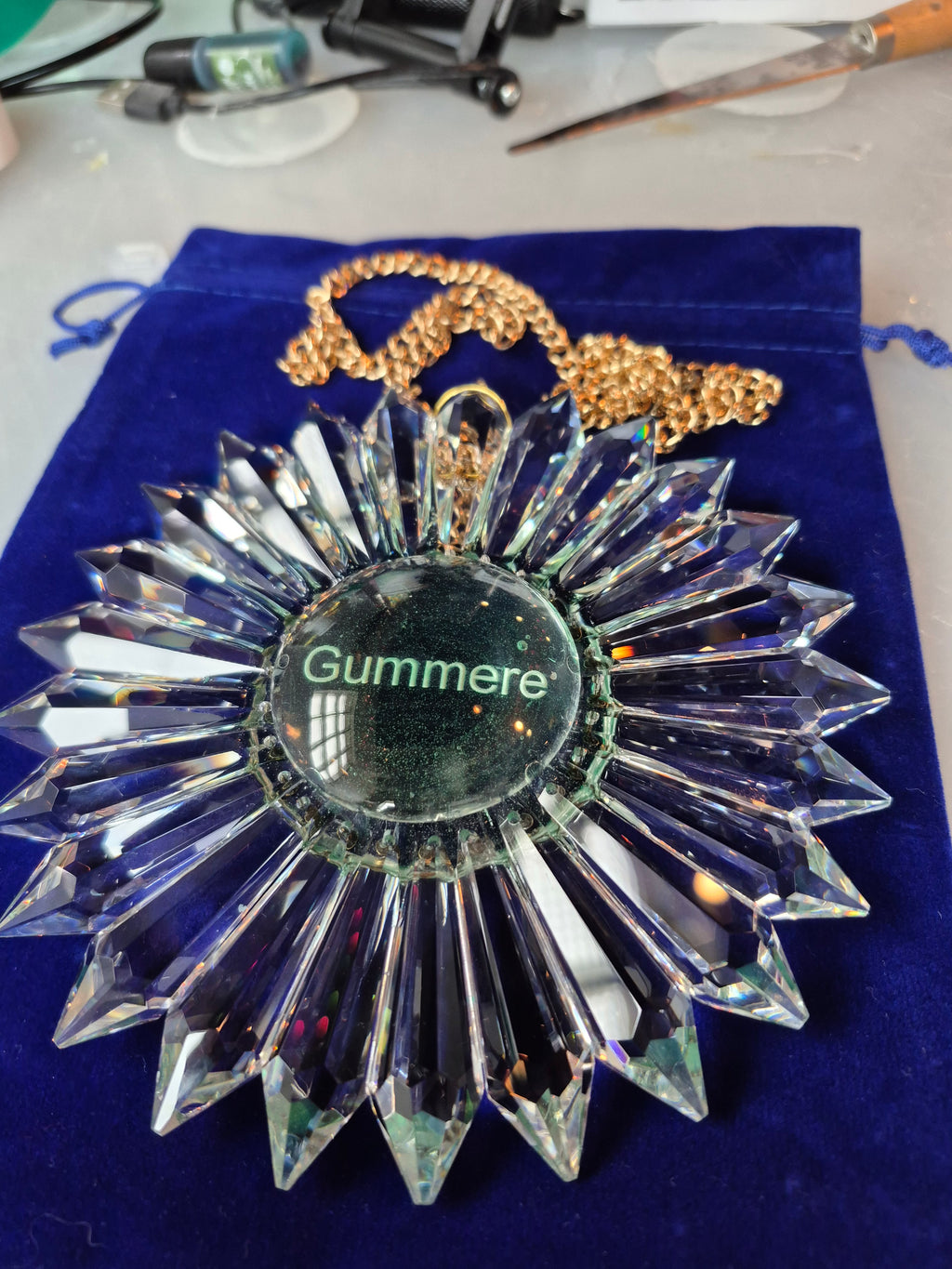 A clear faceted crystal sunburst rainbow maker ornament with a gold-tone chain, displayed on a blue velvet pouch. The center of the pendant features a circular clear insert with the name 'Gummere' printed in green.