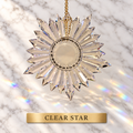 A clear faceted star-shaped lead crystal prism ornament with a gold hanging chain, shown against a white marble background with rainbow light reflections. A gold label at the bottom reads 'CLEAR STAR'.