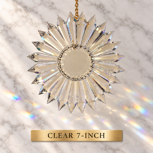 A clear faceted sunburst-style hanging ornament with a circular center and radiating pointed crystal prisms suspended from a gold chain against a white marble background with rainbow light reflections. A gold label at the bottom reads 'CLEAR 7-INCH'.