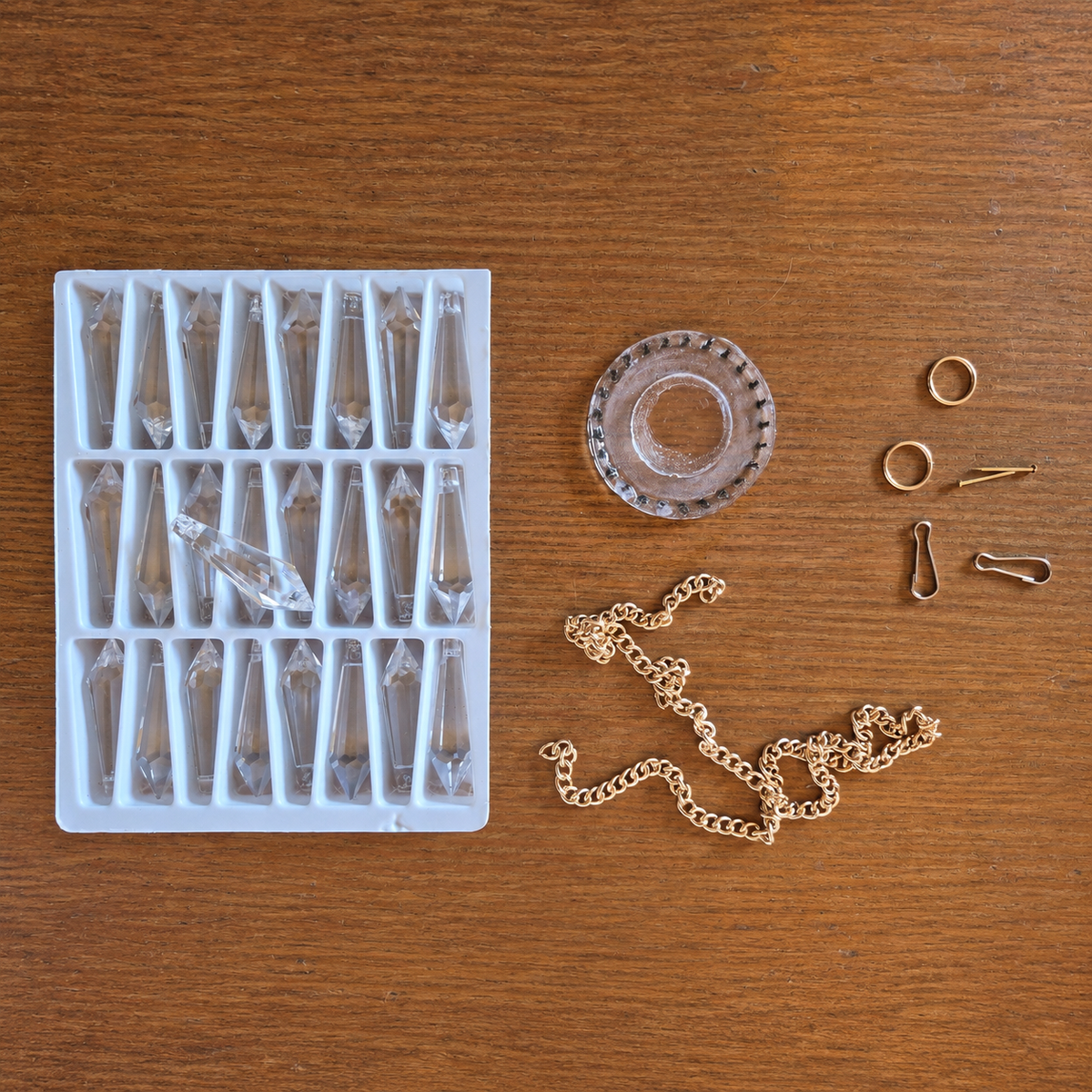 Jewelry making components laid out on a wooden surface, including a white tray of clear faceted crystal beads, a clear glass stopper, gold chain links, rings, findings, and a small metal piece.