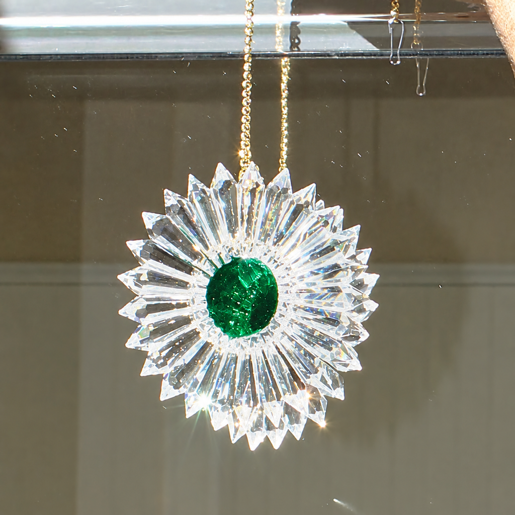 A crystal sunburst ornament with a round green emerald center, hanging from a gold chain against a mirror.