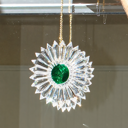 A crystal sunburst ornament with a round green emerald center, hanging from a gold chain against a mirror.