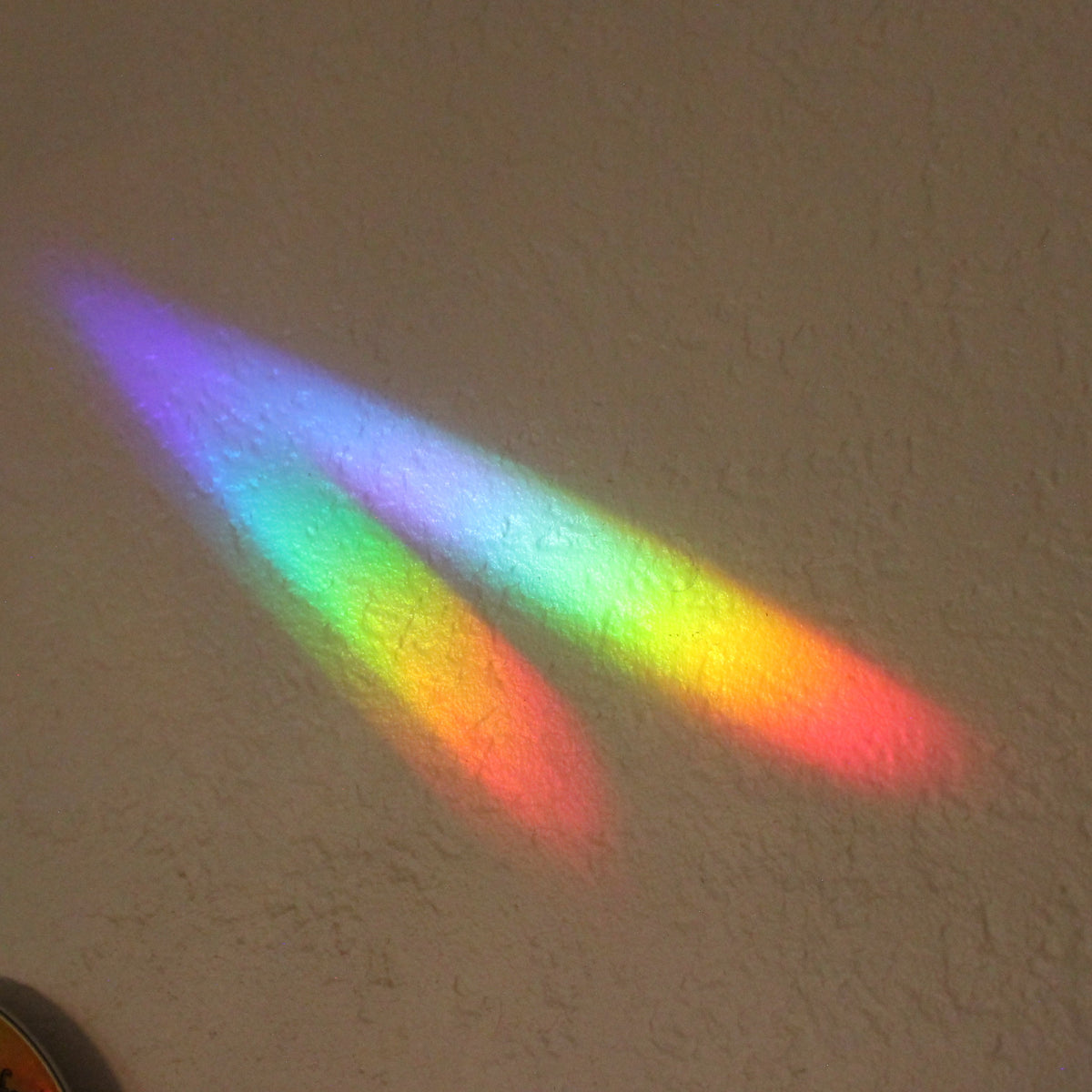 A rainbow light casting of two diagonal beams of colored light lands on a beige textured wall.