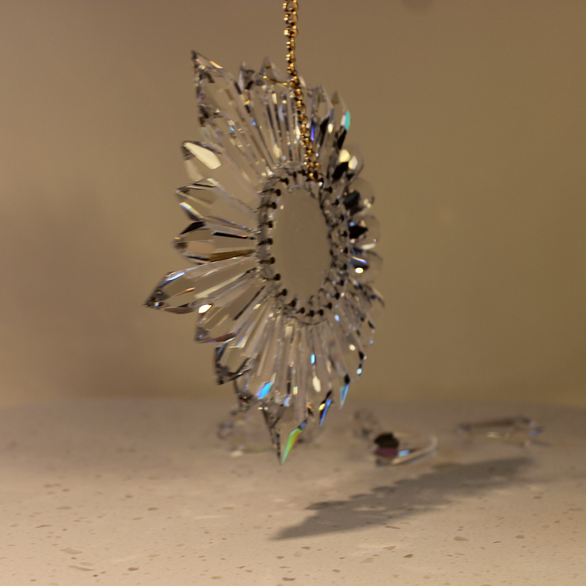 A clear faceted rainbow making prism art with a gold hanging chain, photographed against a warm beige background.