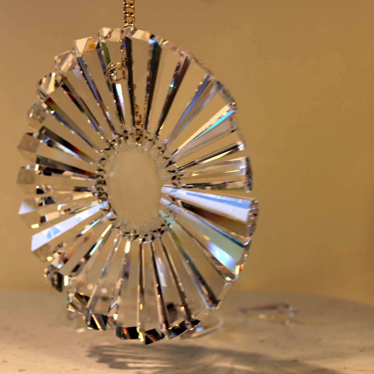 Oblique view of clear lead crystal prisms in radial pattern hanging by a gold chain against a warm beige background