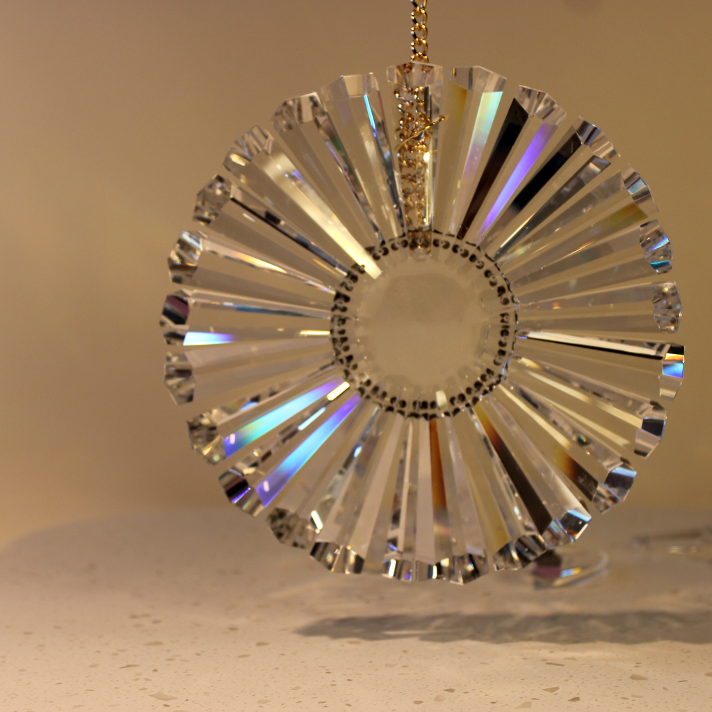 A clear faceted hexagon shaped prisms in a circular ornament with a gold hanging chain, photographed against a warm beige background.