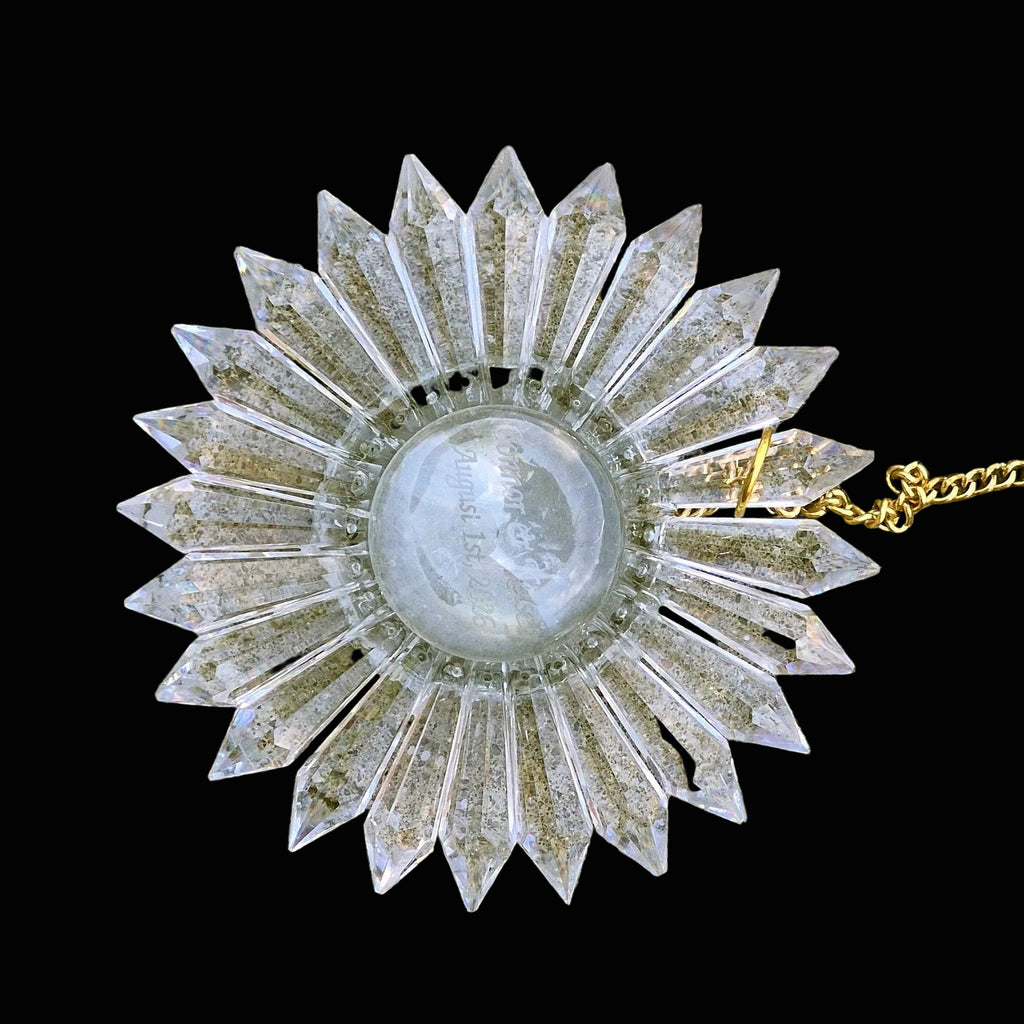A clear faceted crystal sunburst prism suncatcher with a gold chain, displayed on a textured sandy surface.