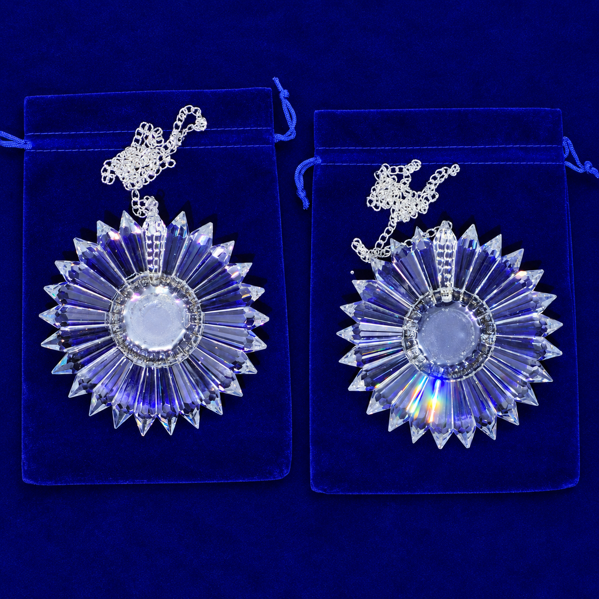 Two matching clear faceted crystal sunburst pendants with silver chains, displayed on blue velvet pouches against a dark blue background.