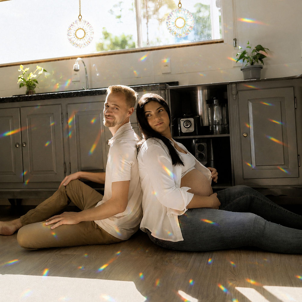 A couple expecting a baby sit on the kitchen floor enjoying each other and the rainbow spectrums coming from the two prism suncatchers hanging in the sunny window.