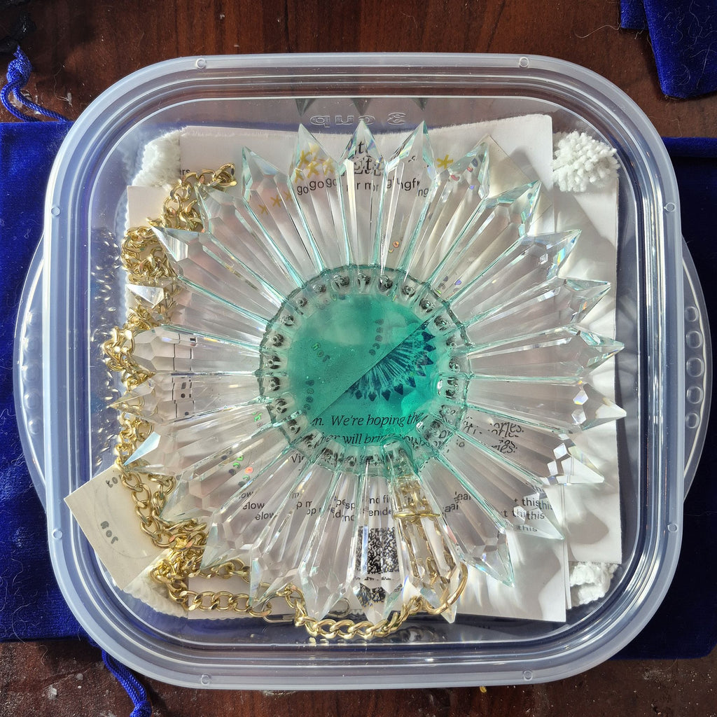 A clear faceted starburst keepsake box containing a turquoise green circular center piece with printed text, gold chain accents, and white paper inserts, photographed from above on a dark wooden surface with blue fabric around it.