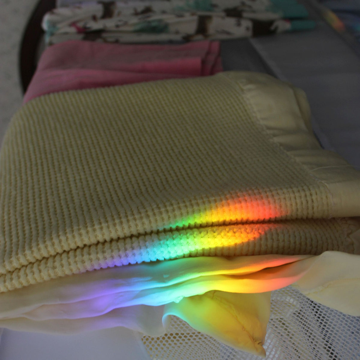 A folded cream-colored textured blanket or throw with a soft white mesh underside, showing a rainbow of reflected light across the center. Other folded blankets in pink and patterned fabric are stacked in the background.