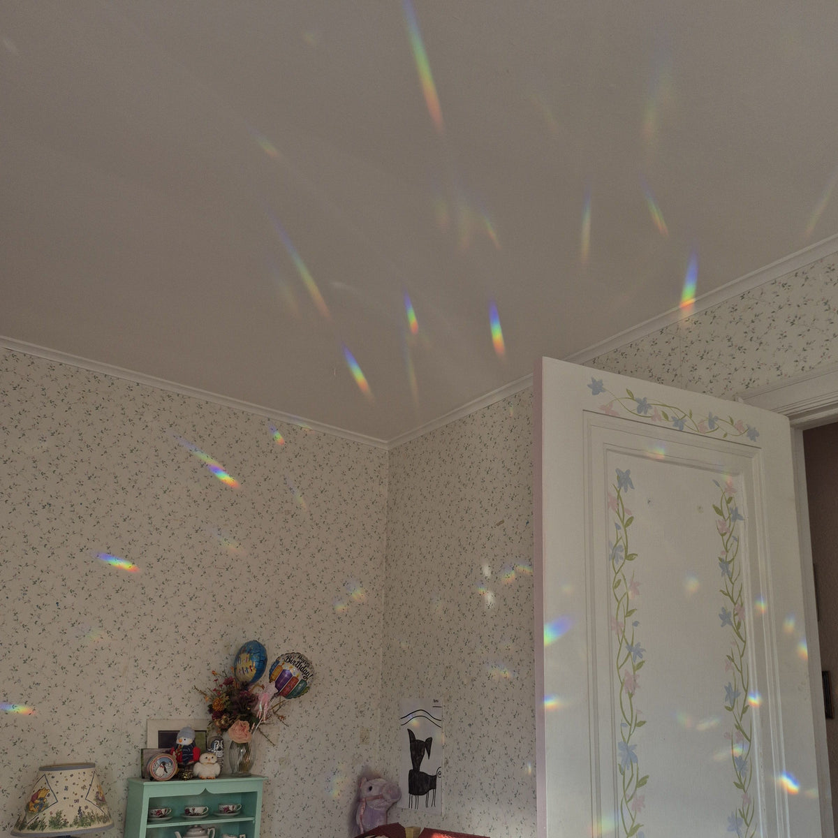 prism suncatcher in the window casting rainbow light beams across a decorated room with floral wallpaper, a white door, and a small shelf.