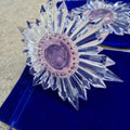 glass bubble disk over transparent pink center of two star-shaped prism suncatchers resting on blue pouches in the daylight