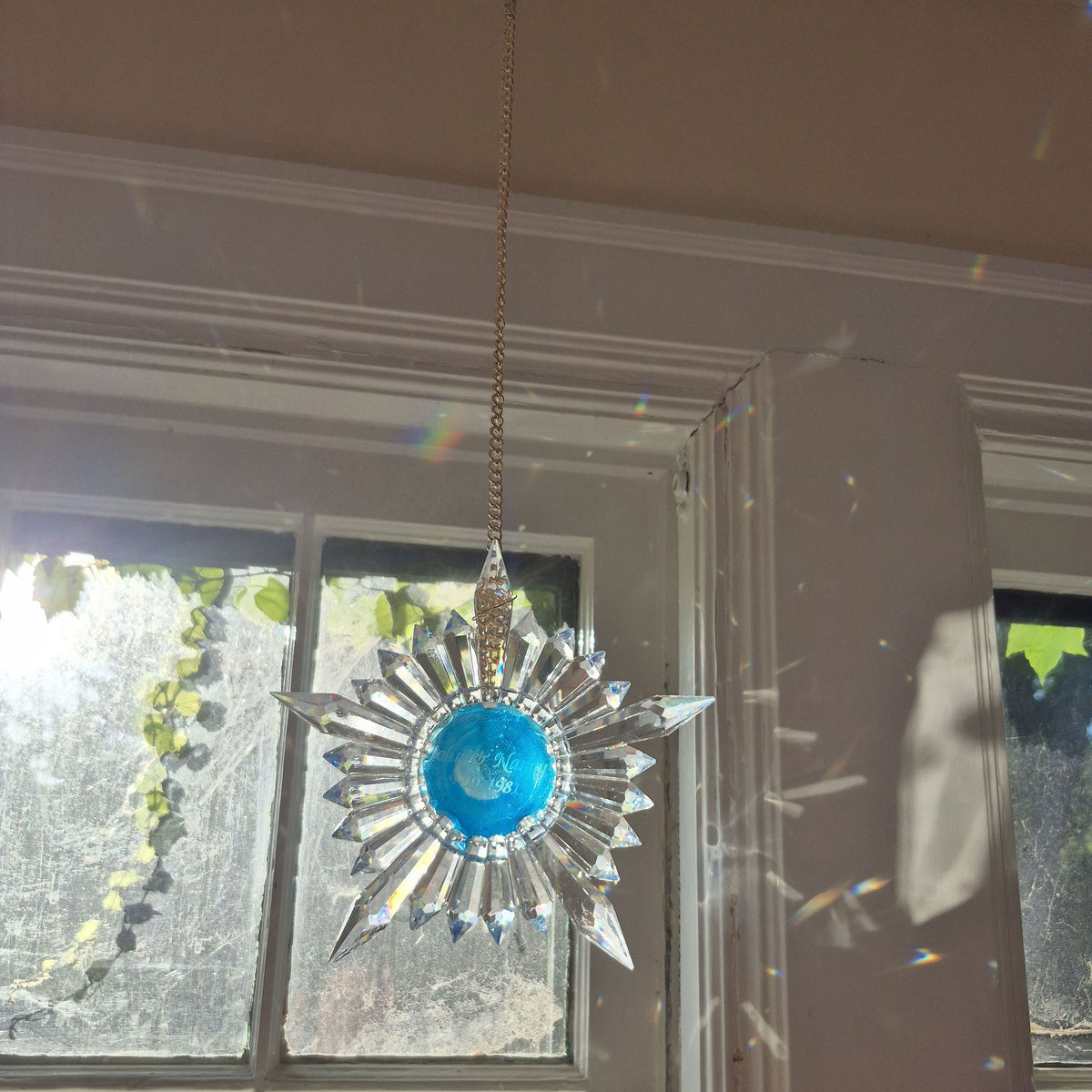 A clear faceted star-shaped suncatcher with a bright blue center, hanging in front of a window with sunlight and rainbow reflections.