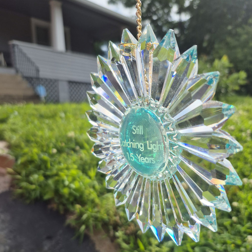 A faceted clear glass sunburst ornament with a gold hanging chain and a central round green-blue section that reads 'Still Catching Light 15 Years,' photographed outdoors in front of a blurred house and greenery.