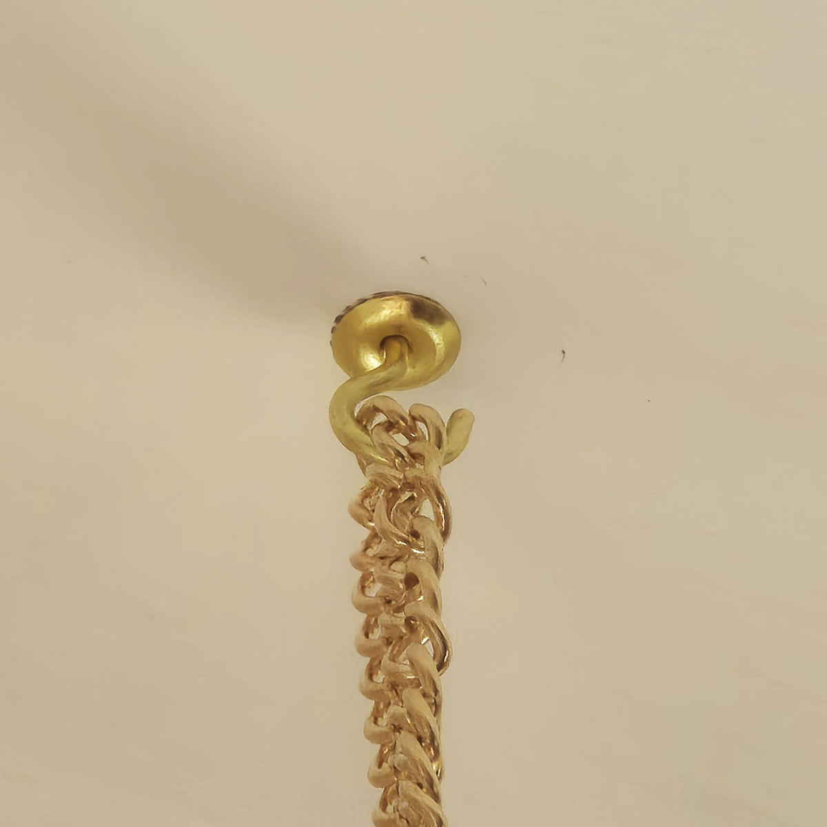 A close-up of a gold-toned chain hanging from a matching gold-toned wall hook or ceiling mount against a plain off-white background.