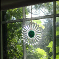 A clear glass sun catcher suncatcher with a deep green center and radiating starburst points, hanging in a window with green foliage visible outside.
