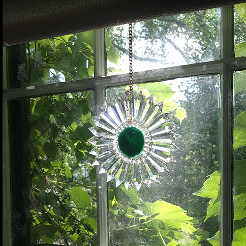 A clear glass sun catcher suncatcher with a deep green center and radiating starburst points, hanging in a window with green foliage visible outside.