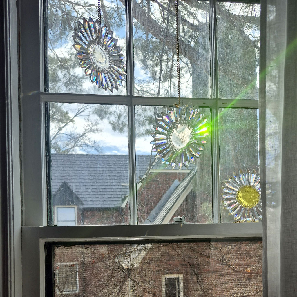 Three circular stained glass sunburst ornaments with hanging chains are displayed on a multi-paned window, with outdoor trees, a brick house, and a bright sunbeam visible through the glass.