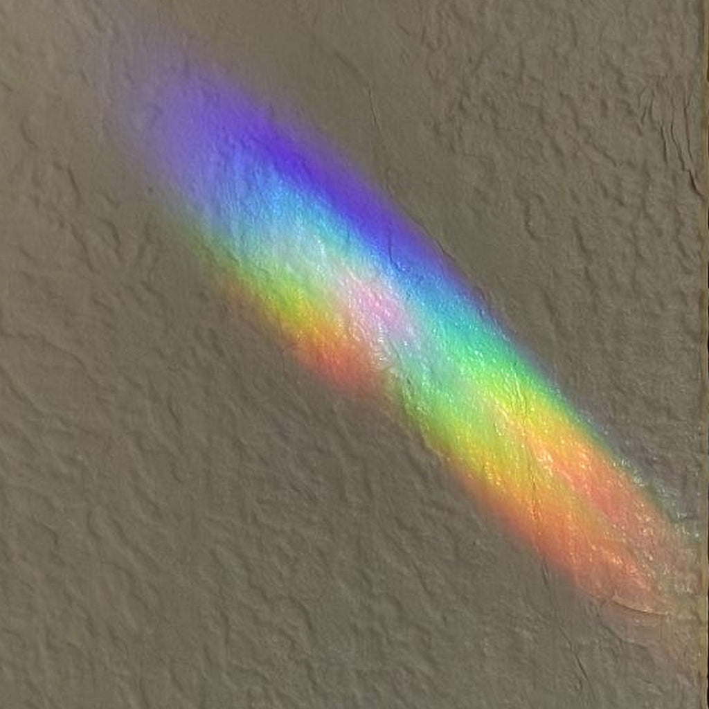 Two overlapping diagonal rainbow light beams on a textured beige wall, showing a spectrum of mixing colors including unusual shades of purple, blue, green, yellow, and orange.