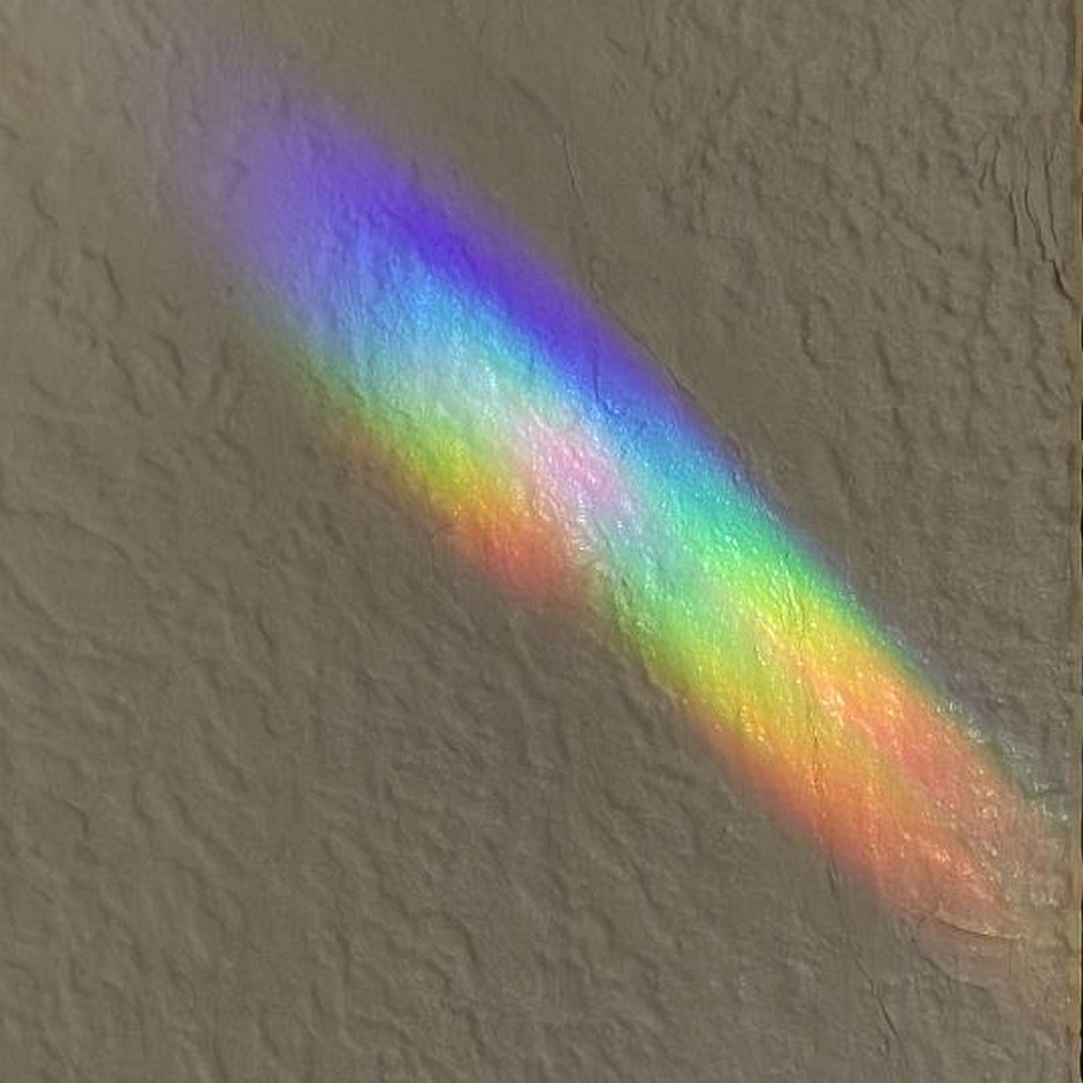 Two overlapping diagonal rainbow light beams on a textured beige wall, showing a spectrum of mixing colors including unusual shades of purple, blue, green, yellow, and orange.