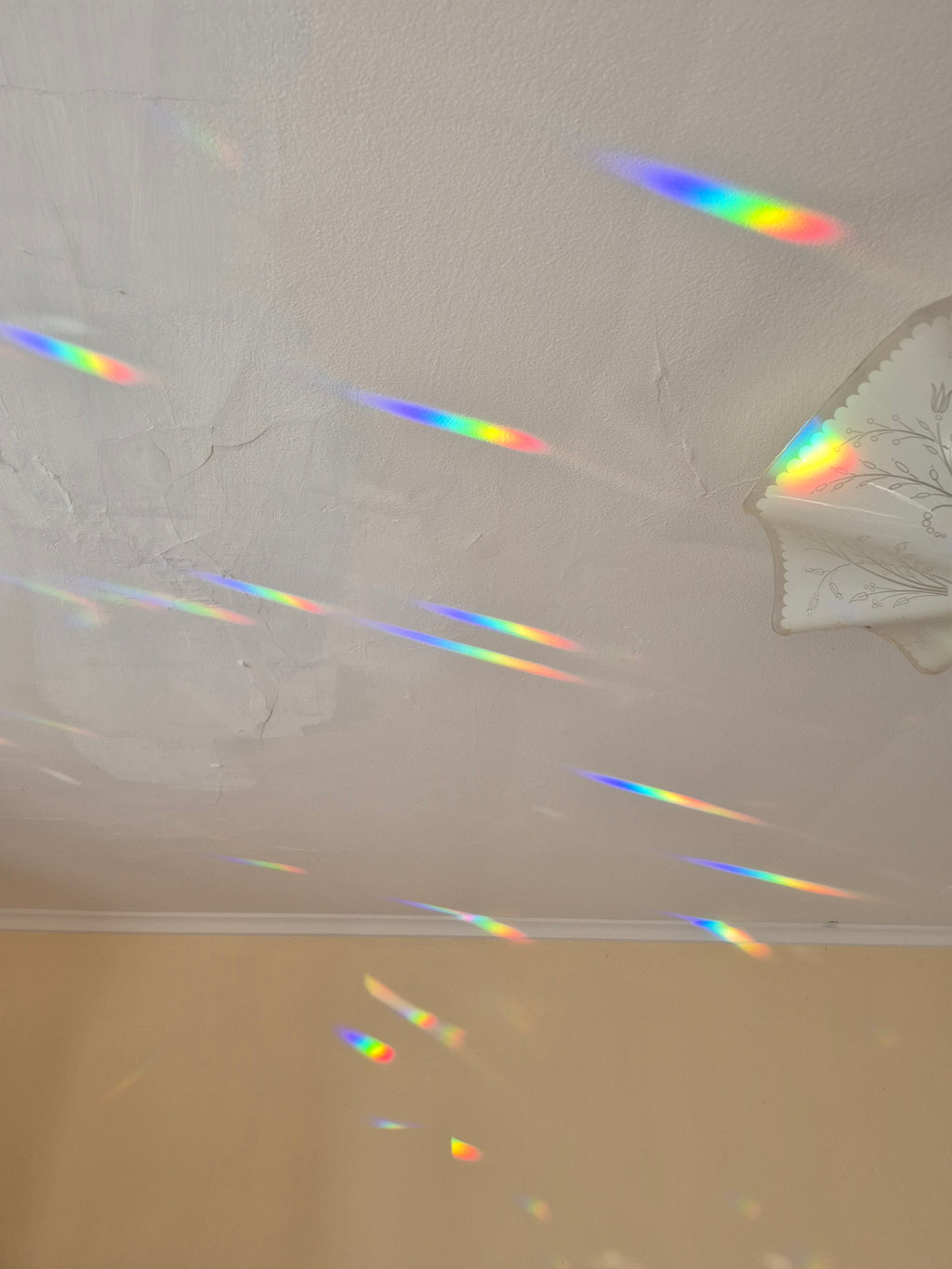 A white textured ceiling with multiple colorful rainbow light streaks and prisms scattered across it, with a beige wall visible at the bottom.