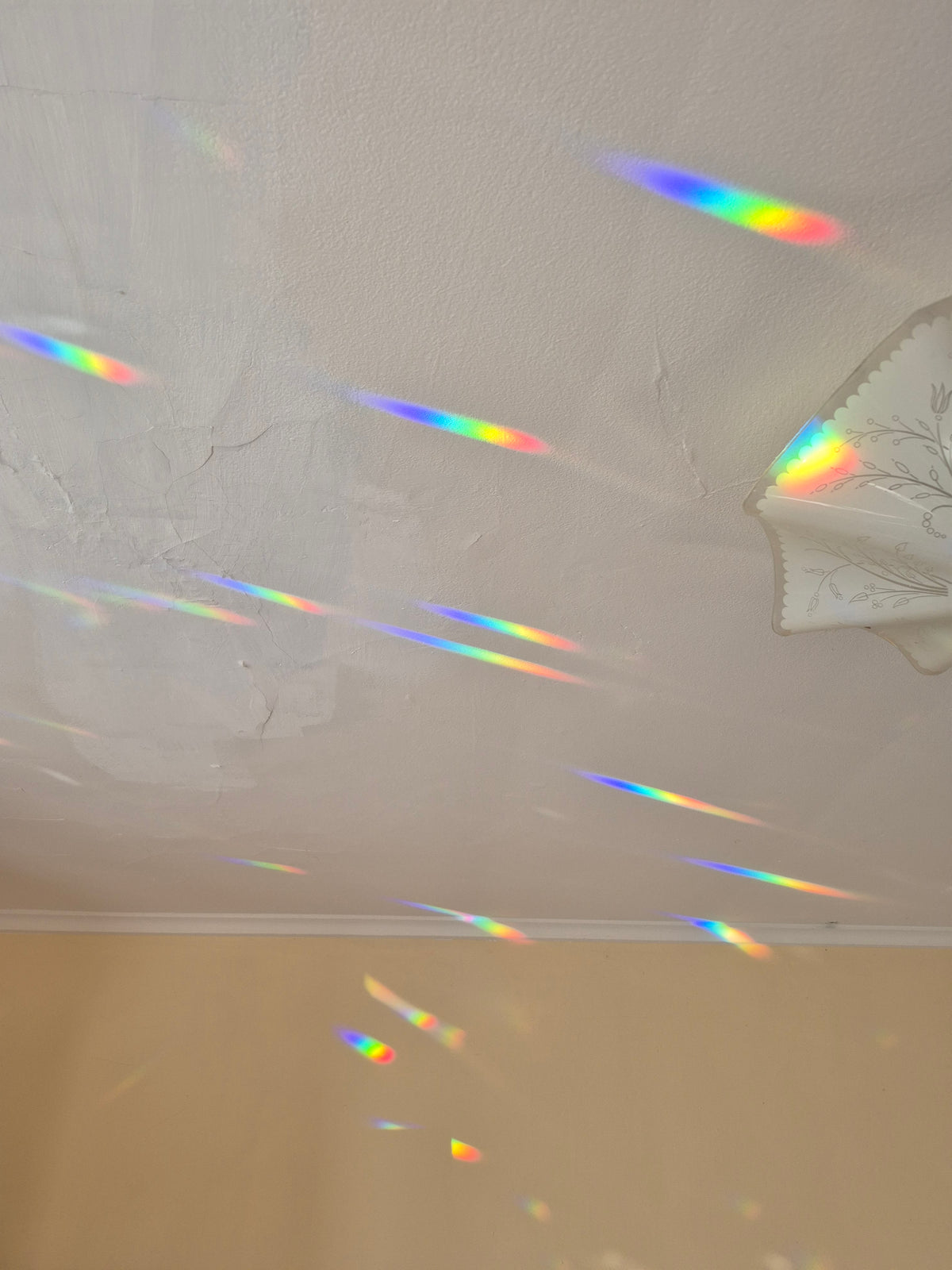 A white textured ceiling with multiple colorful rainbow light streaks and prisms scattered across it, with a beige wall visible at the bottom.