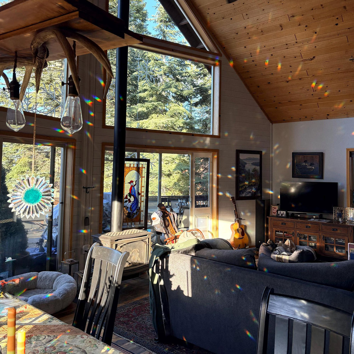 A cozy, sunlit living room inside a rustic wooden chalet with large windows, showing a gray sofa, wooden ceiling, and natural light filtering through. A rainbow prism effect is visible across the room from light beams outside.