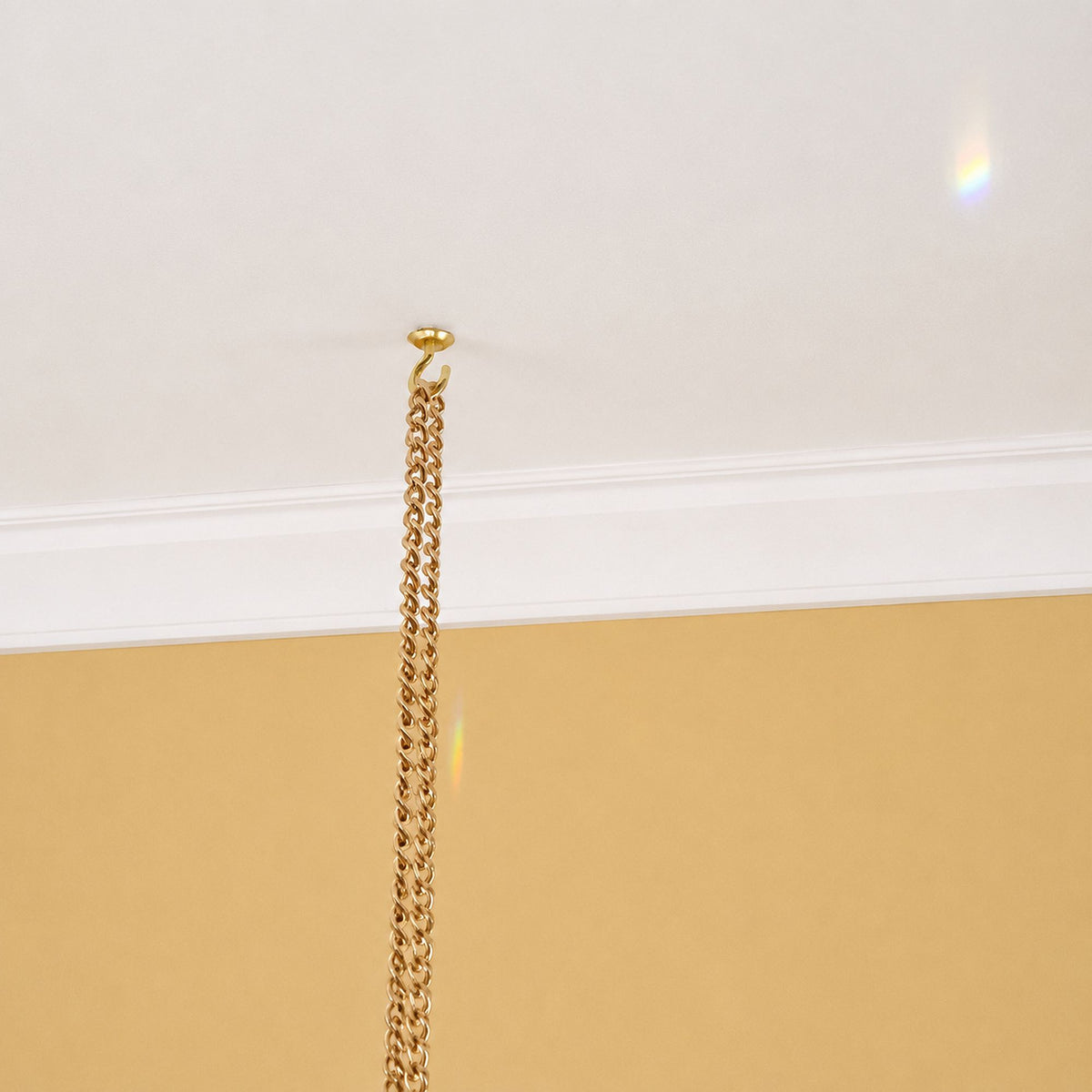 A close-up of a gold metal chain hanging from a ceiling hook, positioned below white crown molding against a beige wall.