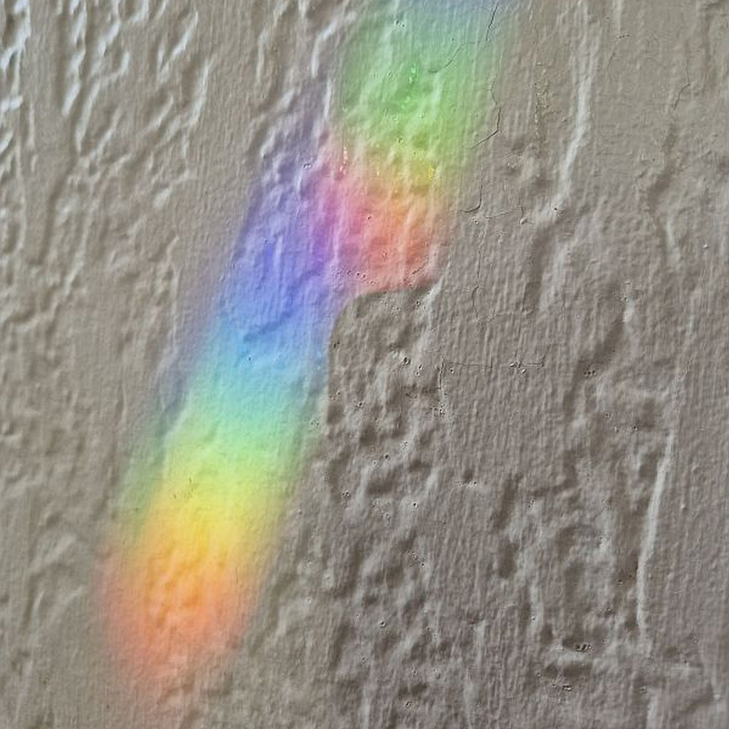 A textured gray plaster wall with a diagonal rainbow light beam reflecting off it, showing a spectrum of pastel like colors from red and orange to green, blue, and purple.