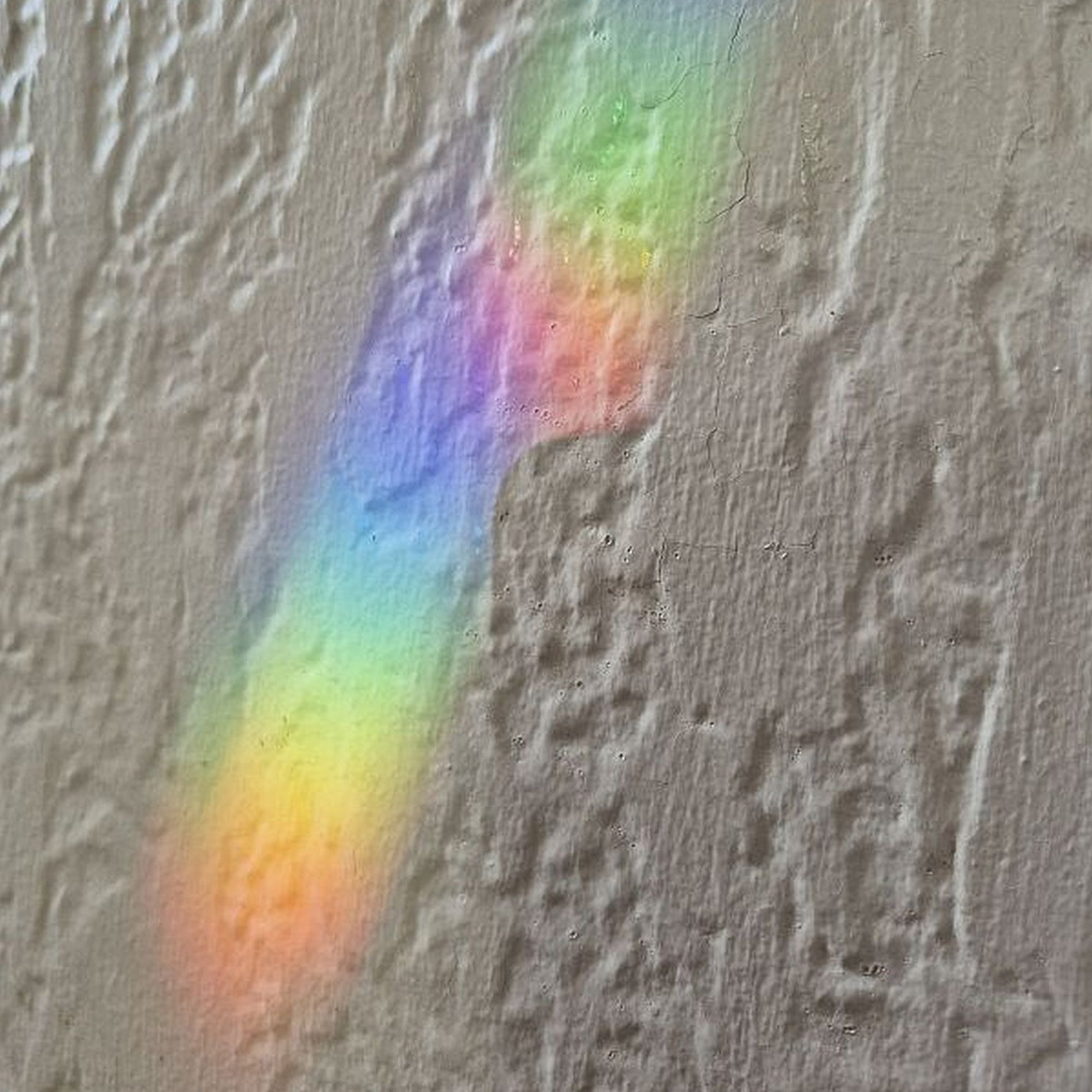 A textured gray plaster wall with a diagonal rainbow light beam reflecting off it, showing a spectrum of pastel like colors from red and orange to green, blue, and purple.