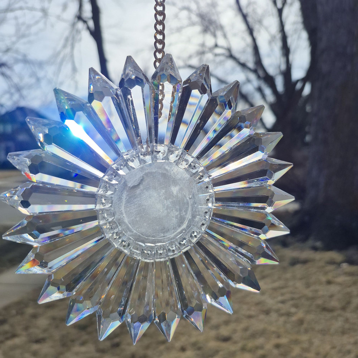 A clear faceted crystal sun gazing ornament with a starburst design, hanging from a metal chain against an outdoor background of trees and grass.