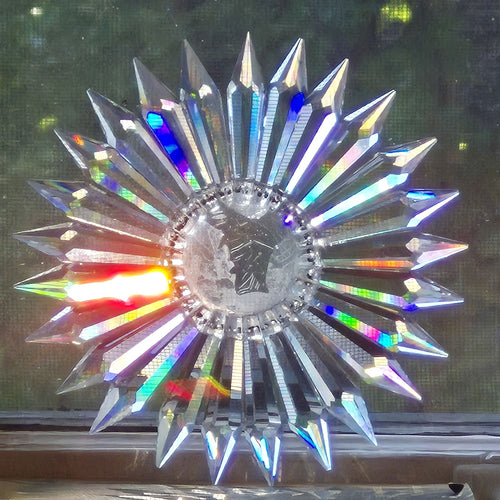A faceted starburst window ornament made of Egyptian lead crystal prisms having 25 sharp rays radiating from a circular center, shown against a dark window background.