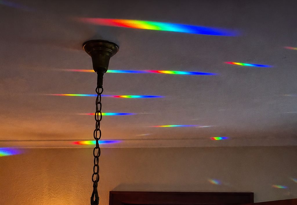 A hanging light fixture with a dark metal chain and canopy, shown in a room with rainbow-colored light beams projected across a ceiling and wall.