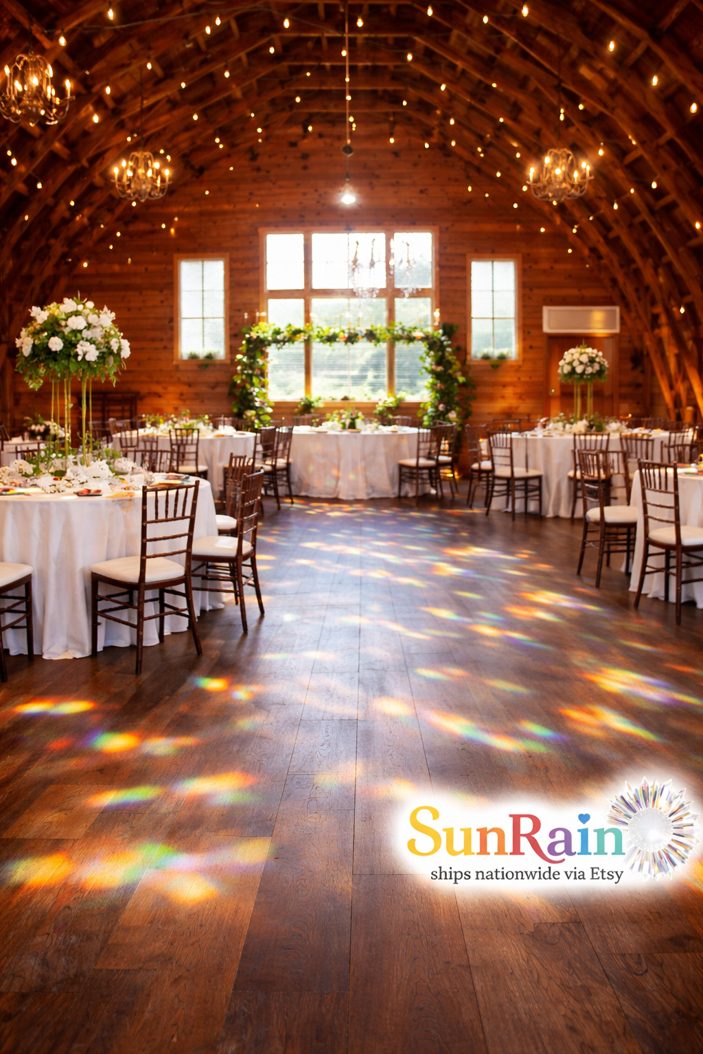 A rustic barn event space with wooden arched ceiling, string lights, chandeliers, and tables set with white linens and floral centerpieces on a wood floor with rainbow light projections from the sunny window