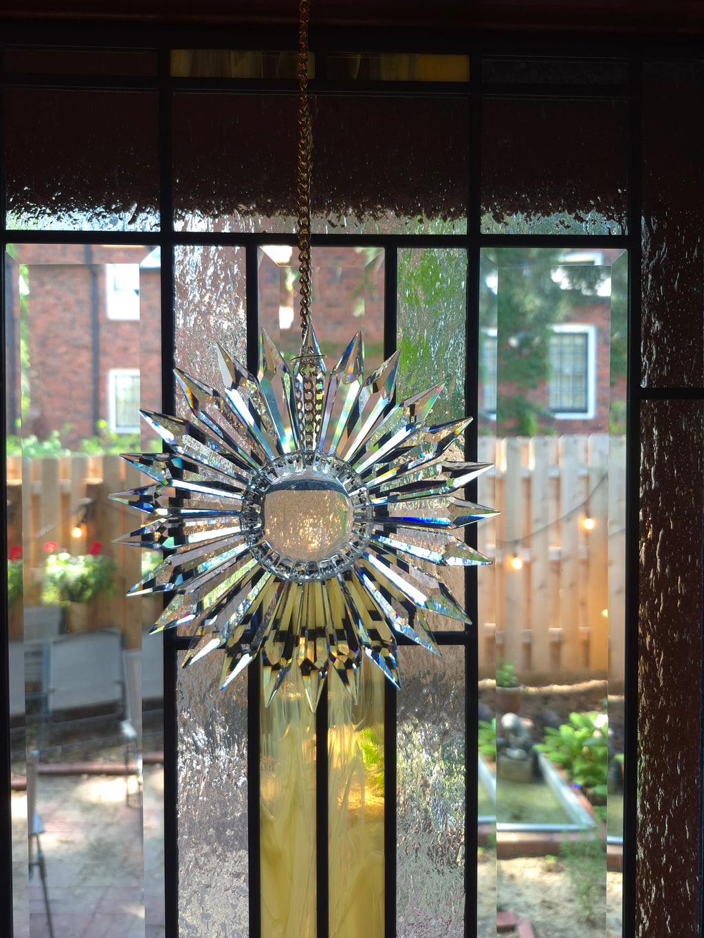 A vintage-style starburst sunburst stained glass window ornament hanging in front of a multi-pane window, with a backyard garden visible in the background.
