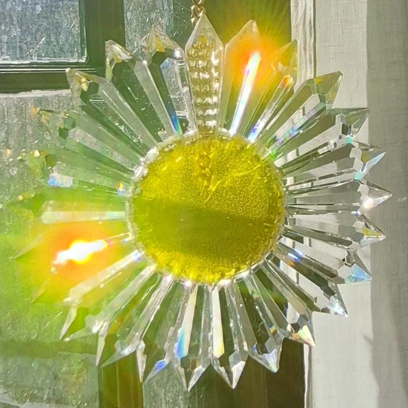 A clear glass sunburst suncatcher with a yellow-gold center, reflecting sunlight in bright rainbow prisms against a window background.