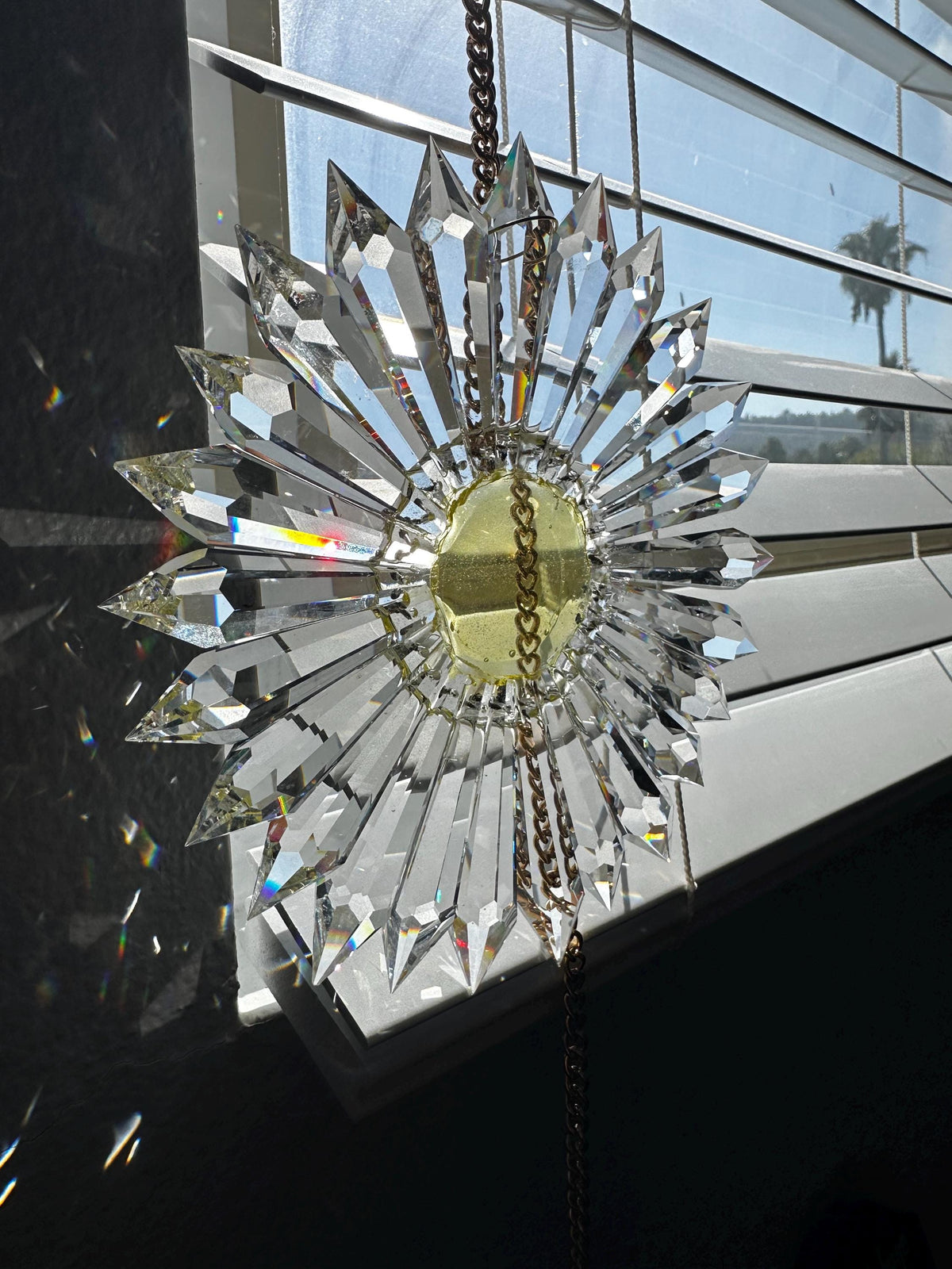 Customer photo of 7-inch crystal sun catcher in a starburst design, featuring a central gold-toned metal ring and hanging chains, shown hanging in front of a window.