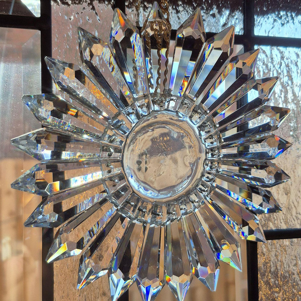 A clear crystal prism suncatcher in a radial pattern hanging from a chain, featuring multiple faceted star-shaped prisms radiating from a round central base over Art Deco divided light window