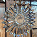 A clear crystal prism suncatcher in a radial pattern hanging from a chain, featuring multiple faceted star-shaped prisms radiating from a round central base over Art Deco divided light window